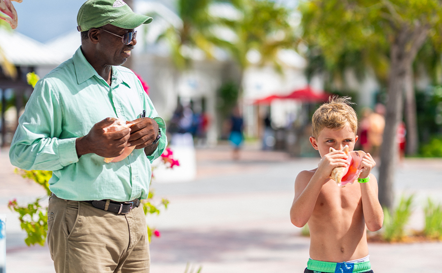 A man in a green shirt instructing a young boy on how to blow a conch shell, with palm trees and a bright outdoor setting in the background.