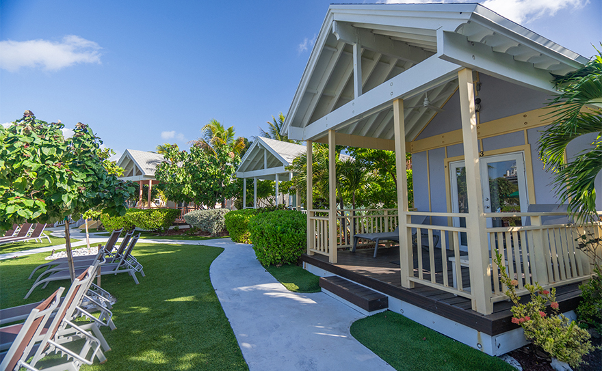 A row of cozy beach cottages with white roofs surrounded by lush greenery and palm trees, under a bright blue sky.